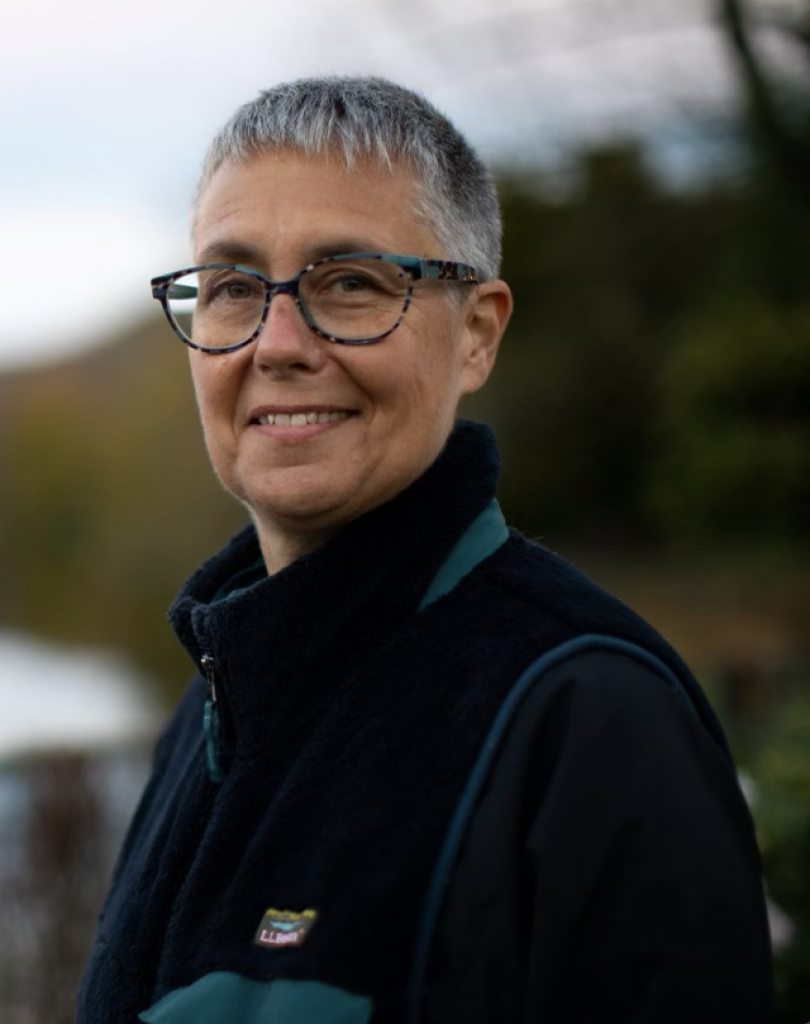 a square photo of Steph, a woman with very short hair, wearing glasses and a dark blue vest