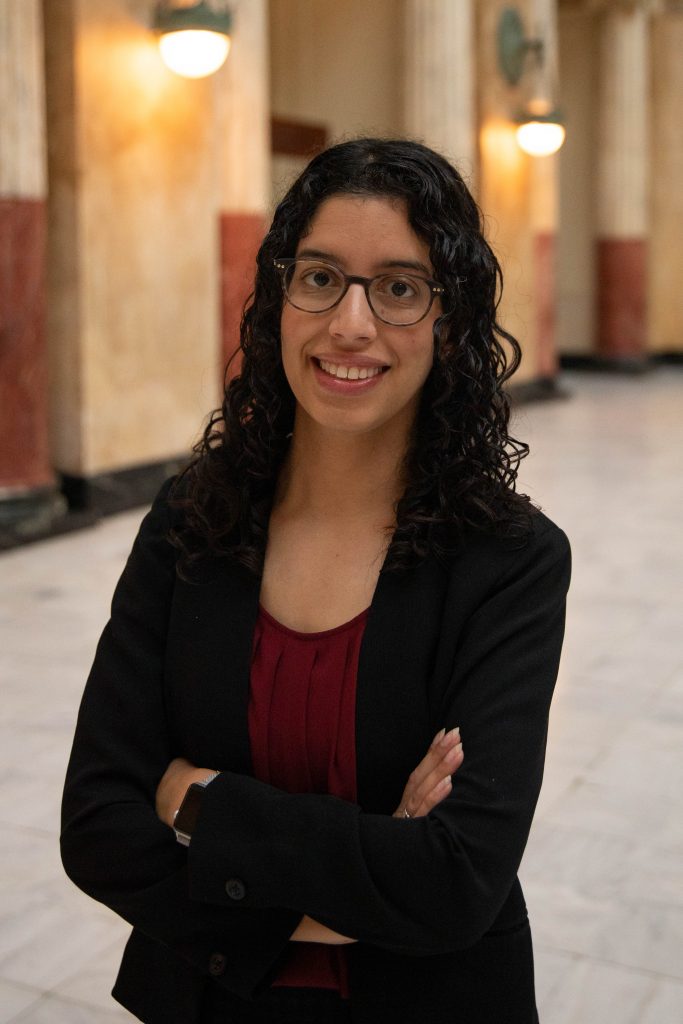 Photo of Zainab Alkebsi, a Brown woman with curly, dark hair and glasses in a purple top and black jacket.