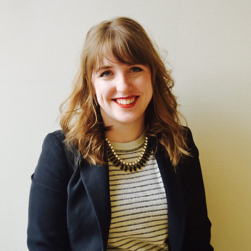 Photo of Ali Szemanski, a woman with wavy brown hair and blue eyes in a grey and white striped top and dark blue jacket.